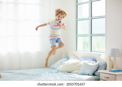 Child Jumping On Bed In White Bedroom With Big Window. Kids Play At Home. Fun Jump On Parents Bed. Room And Interior Design. Boy At Home. Sunny Morning, Weekend Family Relaxing Time.