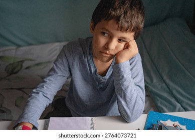 A child leaning on a table while doing homework, showing an expression of tiredness or lack of motivation, in a home setting; ideal for concepts related to education, aca-demic effort, routine, and children’s emotions.