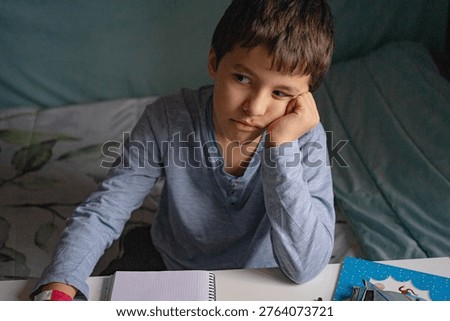 A child leaning on a table while doing homework, showing an expression of tiredness or lack of motivation, in a home setting; ideal for concepts related to education, aca-demic effort, routine, and children’s emotions.