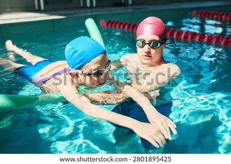 Child learning swimming with instructor in pool, practicing floating and water safety skills during lesson. Useful for swim school promotion, kids fitness, healthy lifestyle marketing