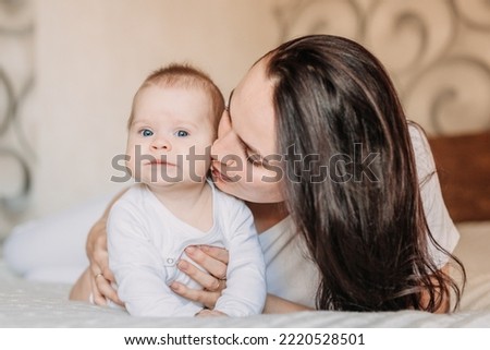 child lies on the bed and looks into the camera. Beautiful cute baby next to mom, gently hugging and kissing.