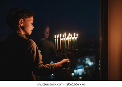 The child lights the menorah for Hanukkah on the windowsill. The boy in the kippah sitting by the window. Jewish holiday. Tradition is a religious ritual. Sunset. The first star. Judaism