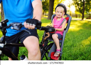 child a little girl in a Bicycle helmet smiles sitting in a child's Bicycle seat against the background of a Park and green grass.