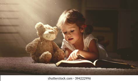 child little girl reading a magic book in the dark home with a toy teddy bear