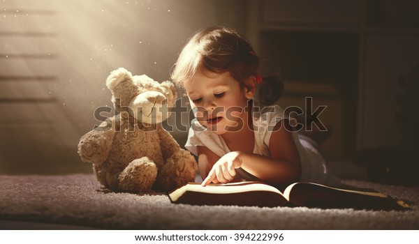 おもちゃのテディベアで暗い家の中で魔法の本を読む少女 の写真素材 今すぐ編集