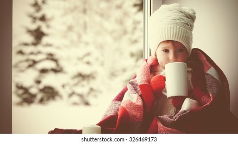 Child Little Girl Sitting By The Window With A Cup Of Hot Tea And Looking At The Winter Forest