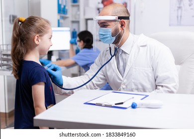 Child looking at doctor while he's listening heart with stethoscope. Health pediatrician specialist providing health care services consultations treatment in protective equipment for consultation