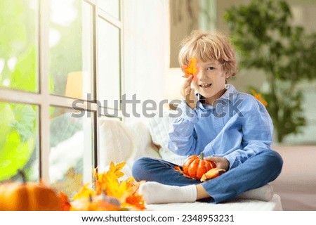 Child looking out of the window. Autumn cozy family home decoration. Kids at home in fall watching golden leaves in sunny garden. Little boy with pumpkin on Thanksgiving day or Halloween.