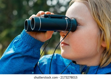 Child Looking Through Binoculars In The Park. Closeup Portrait Of A Boy Exploring Wildlife. Bird Watching, Hiking, And Adventure Concept.