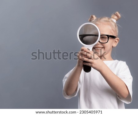 child looking through magnifying glass on gray background. Portrait of curious smiling little girl holding magnifier and exploring world. Investigation, discovery and vision concept. Copy space