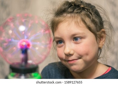 The Child Looks At The Plasma Ball. Portrait Of A Little Girl With A Magic Ball. Kid Plays Witchcraft And Magic. Selective Focus