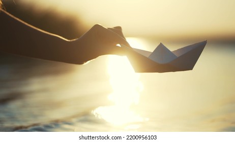 The Child Lowers The Paper Boat Into The Water. Happy Family Fantasy Child Dream Concept. A Child Plays With A Paper Boat Ship. Child's Hand Launches A Boat In The Park In The Pond Lifestyle