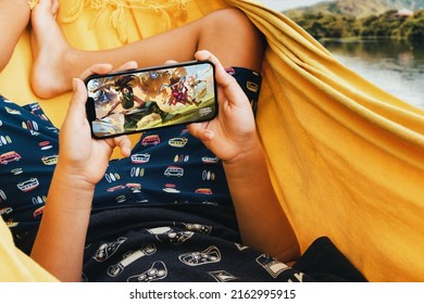 Child lying in hammock with League of Legends Wild Rift LOL mobile game app on smartphone screen. Lake in the background. Rio de Janeiro, RJ, Brazil. May 2022.