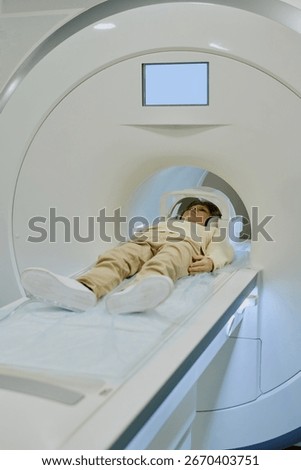 Child lying inside MRI scanner undergoing medical imaging procedure in hospital setting, head supported by cushion, machine surrounding upper body, eyes closed, hands resting at sides