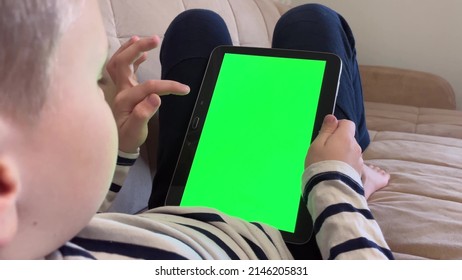 Child Lying On The Couch And Using A Digital Tablet PC With Green Screen, Back View. Boy Holding A Tablet With Green Screen In Hand