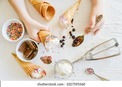 The child Makes his own Ice cream: Top View, Children's Hands Holding a cone of ice Cream On the table Chocolate, Spoon, Topping, Berries, Sprinkles, Jam.