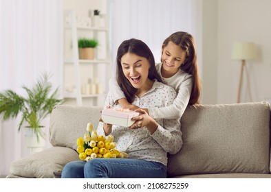 Child makes wonderful surprise for mom on Mother's Day. Happy Caucasian woman gets gift and bouquet of fresh flowers from daughter. Excited smiling mommy sitting on sofa and looking at decorated box - Powered by Shutterstock