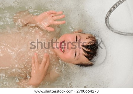 Child making bath time fun, looking at camera and smiling. Playful bubbled water in bathtub with a shower hose. Baby hygiene and skin care. Child aged 4 years (four years old)