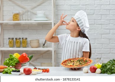 Child Making Tasty Delicious Gesture By Kissing Fingers. Little Girl In Chef Hat And An Apron Cooking Pizza In The Kitchen.