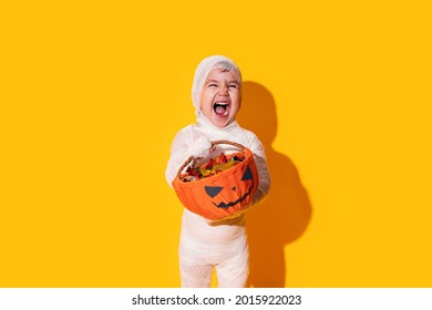 Child in mummy costume holding basket of chocolates in front of yellow background. - Shutterstock ID 2015922023