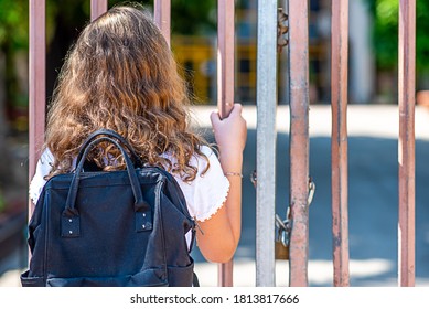 Child Near The Gate Of The Closed School. Back View Blonde Teenage Girl Outside A Closed School Fence.