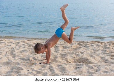 A Child On The Beach Does Sports And Performs Gymnastic Exercises Against The Background Of The Sea.  A Child Does A Handstand On The Beach