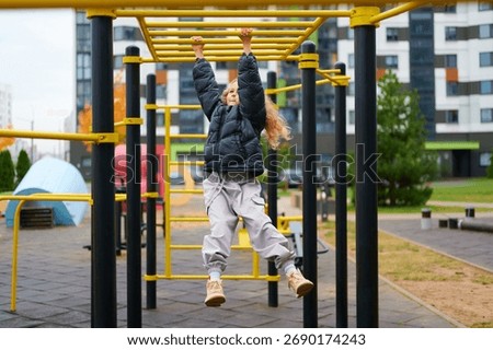 child on monkey bars at playground in urban setting. little girl wearing winter jacket and boots, enjoying active outdoor playtime. bright equipment contrasts with modern buildings