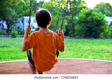 Child On A Playground Swing. A Young Boy Sits On A Playground Swing Alone Looking Into The Distance. View From Behind. Rear View. Feels Sad, Lonely, Lost, Bored.