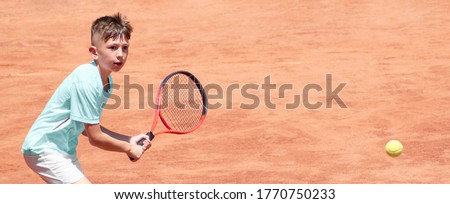 Child on tennis court with a racket in his hands is ready to hit backhand. Boy tennis player in motion and action. Emotions on face of young athlete during sports game. Panoramic. Banner. Copy space