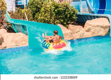 Child On Water Slide At Aquapark During Summer Holiday. Young Boy Or Kid Has Fun Splashing Into Pool