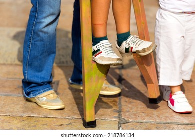 Child On Wooden Stilts / An Adult Helps A Child To Walk On Wooden Stilts, View Of Sun Legs And Feet