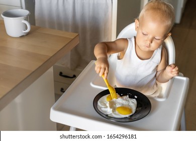 A Child Of One And A Half Years Old Independently Tries To Eat With A Fork While Sitting In A Child Seat In The Kitchen. Fried Fried Eggs For Breakfast For The Baby.