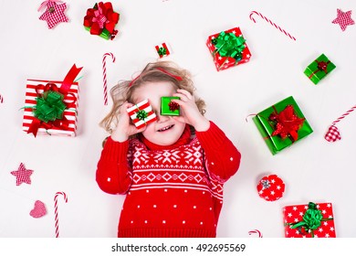 Child Opening Christmas Presents. Little Girl In Knitted Winter Sweater With Present Box. Kids Open Gifts. Toddler Kid On The Floor Under Decorated Xmas Tree. Children Play With Gift Box And Candy.