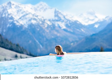 Child In Outdoor Infinity Pool With Snowy Mountain In The Background. Family Vacation In Luxury Alpine Resort. Kids In The Alps Mountains. Hot Tub In Snow. Apres Ski Activity For Kids. Children Swim.