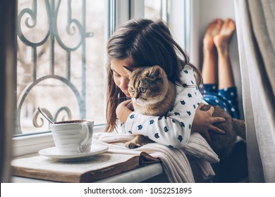 Child In Pajamas Relaxing On A Window Sill With Pet. Lazy Weekend With Cat At Home. Cozy Scene, Hygge Concept.