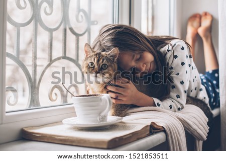 Child in pajamas relaxing on a window sill with pet. Lazy weekend with cat at home. Cozy scene, hygge concept.