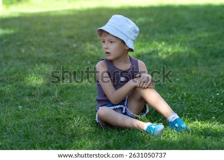 A child in a panama hat sits on the grass in the park. People