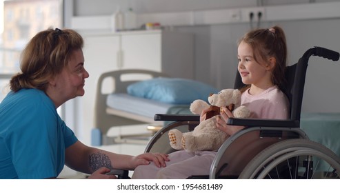 Child Patient In Wheelchair Sitting In Hospital Ward Talking To Female Nurse. Medical Worker Entertaining And Cheering Little Disabled Girl In Wheelchair