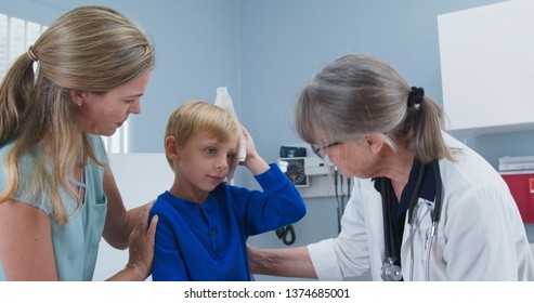 Child At Pediatricians Office Holding Ice Pack On His Head With Mother And Doctor In Room. Little Boy Using Cold Compress In Medical Exam Room Receiving First Aid