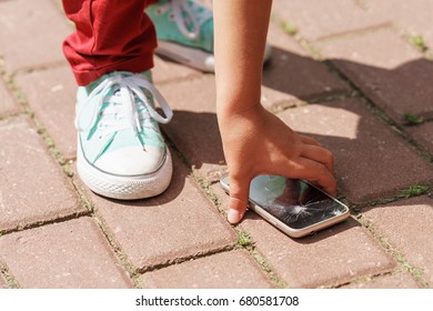 Child Picks Up The Phone With A Broken Screen From The Sidewalk At Day Time