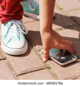 Child Picks Up The Phone With A Broken Screen From The Sidewalk At Day Time