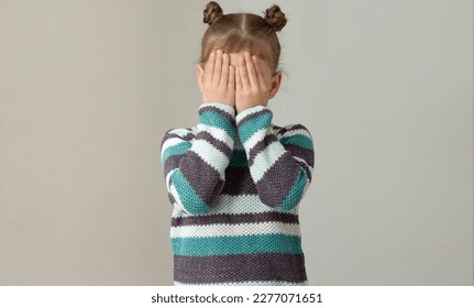 Child placing hands across face to express embarrassment dismay or fear on grey background, caucasian little girl of 6 7 years in green gray striped sweater