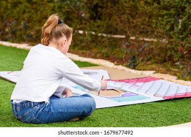 Child Play Outdoors. Back View Teenager Girl Paints A Paper House Template With Colorful Watercolors On The Grass In The Garden. Stress Relief With Art Therapy.