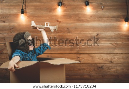 Child is playing in aviator in cardboard box in the room. Little boy having fun against wooden wall and lamps. Imagination and dreams of freedom. Child pilot with airplane. Fast delivery of goods