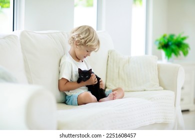 Child playing with baby cat. Kid holding black kitten. Little boy snuggling cute pet animal sitting on white couch in sunny living room at home. Kids play with pets. Children and domestic animals.