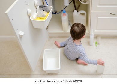 A child is playing with chemical cleaning products under the sink in the kitchen. Baby holds bottles with detergent. Kid aged about two years (one year nine months)