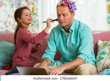 Child playing and disturbing father working remotely from home. Little girl applying makeup with brush. Man sitting on couch with laptop. Family spending time together indoors. 