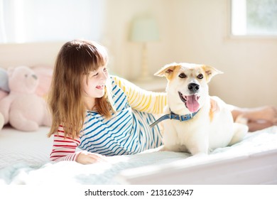 Child Playing With Dog. Girl And Large Dog Relaxing On White Bed At Home. Children And Friendship. Kids Play With Puppy. Kid Hugging Pet. Animal Care. Sunny Morning, Family Time.