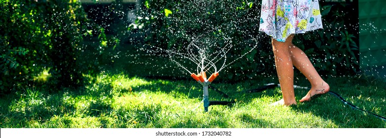 Child Playing With Garden Sprinkler. Happy Girl In Dress Running And Jumping. Kids Gardening. Summer Outdoor Water Fun. Children Play With Gardening Hose Watering Flowers.