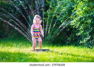 Child Playing With Garden Sprinkler. Kid In Bathing Suit Running And Jumping. Kids Gardening. Summer Outdoor Water Fun. Children Play With Gardening Hose Watering Flowers.